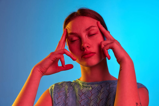Burnout, pain, upset, exhausted. Portrait of pensive girl with closed eyes standing, grabbing head and thinking over blue background in neon light. Concept of health, human emotions, mood, ad - Фото, зображення