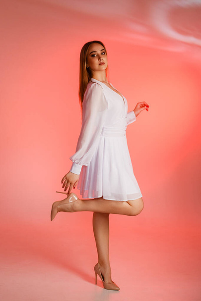 Beautiful girl in a white dress and high-heeled shoes on a pink background. The girl stands with her leg and arms raised, the dress flutters in the wind. - Photo, Image