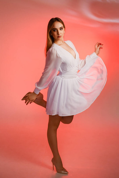 Beautiful girl in a white dress and high-heeled shoes on a pink background. The girl stands with her leg and arms raised, the dress flutters in the wind. - Фото, изображение