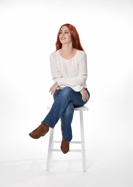 Ganzkörperporträt der schönen Frau Modell mit langen roten Haaren, trägt legeres Outfit weiße Bluse Top und Jeans, isoliert auf weißem Studiohintergrund. Entspannte Sitzhaltung, auf Stuhl sitzend mit gestischer Handhaltung. - Foto, Bild