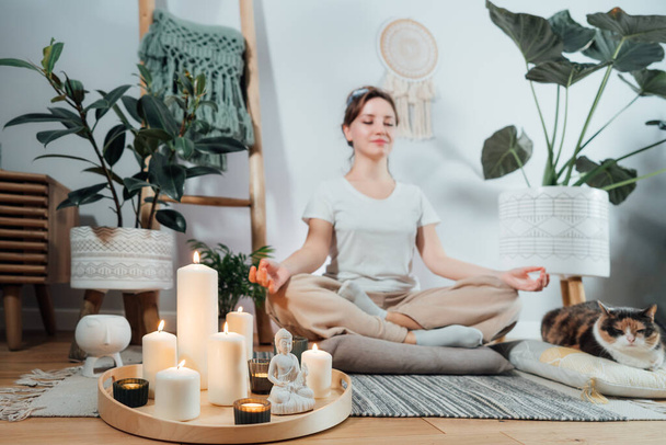 Focus on burning candles and white Buddha statuette on tray with background of woman practicing meditation at home with cat in modern Scandi interior. Zen Composition for yoga practice, relaxation - Foto, imagen