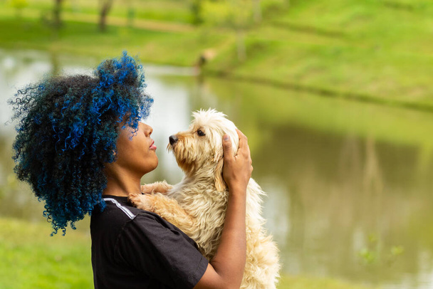Een jonge zwarte vrouw met geverfd blauw haar, met haar hond op schoot, in een goed bebost park, wazig op de achtergrond. - Foto, afbeelding