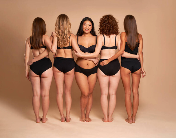 Vista trasera y completa de las mujeres en ropa interior negra de pie sobre fondo desnudo - Foto, Imagen