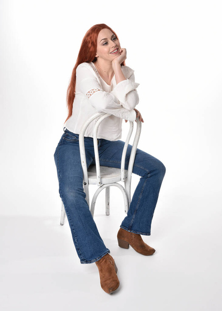 Uzun kızıl saçlı güzel bir kadın modelin tam boy portresi, beyaz bluz ve kot pantolon giyiyor, beyaz stüdyo arka planında izole edilmiş. Rahat bir poz, el hareketleriyle sandalyeye oturmak.. - Fotoğraf, Görsel