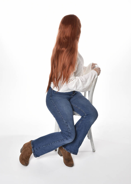 full length portrait of beautiful woman model with long red hair, wearing casual outfit white blouse  top and denim jeans, isolated on white studio background. Relaxed seated pose, sitting on chair with gestural hand poses. - Zdjęcie, obraz