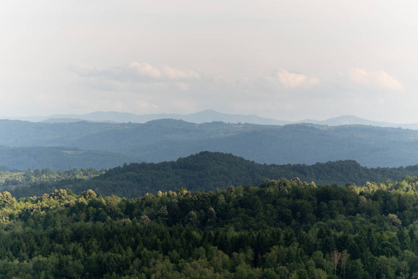 Hügelige Landschaft mit üppigen Wäldern und Wolkenschatten, in der Ferne Bergschichten im Luftdunst - Foto, Bild