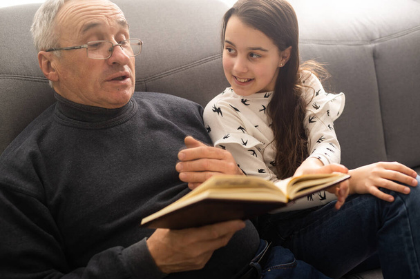 Фото старого дідуся маленька красива онучка сидить диван обіймає будинок карантинної безпеки читати цікаву книгу казка сучасний інтер'єр вітальні в приміщенні
 - Фото, зображення