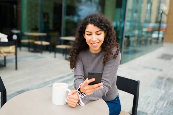 Счастливая молодая женщина улыбается, когда пишет смс на смартфоне или пользуется социальными сетями, наслаждаясь кофе - Фото, изображение