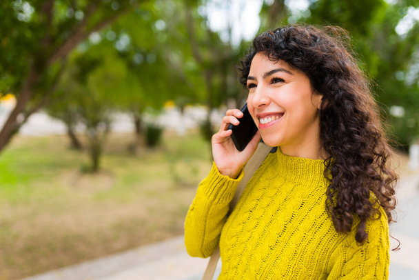 Iloinen nainen hänen 20s kanssa kihara tukka hymyillen puhuessaan hänen älypuhelin ja käyttämällä mobiiliteknologiaa ulkona  - Valokuva, kuva