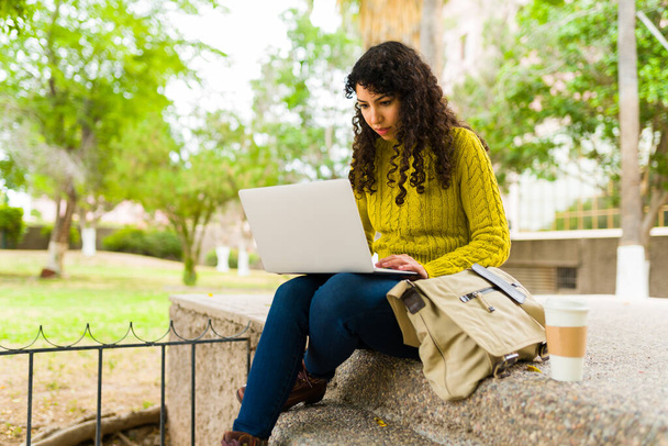 Mujer joven caucásica haciendo trabajo remoto al aire libre en el parque y trabajando en su computadora portátil y una bolsa de mensajero - Foto, imagen