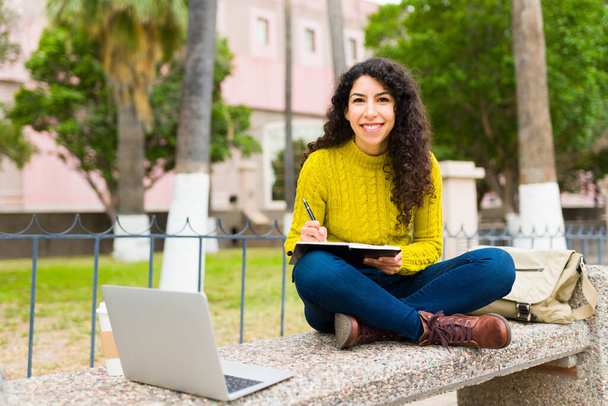 ノートやノートパソコンを使って屋外の公園のベンチに座ってリモートワークをしながら笑顔の美しい若い女性 - 写真・画像