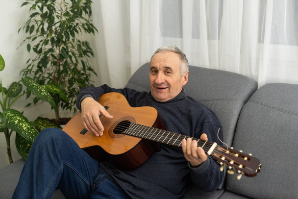 Senior man is playing guitar. Elderly man sitting on the sofa and playing guitar. Portrait of a gray-haired mature man in a sweater learning to play. Enjoying retirement life at home - Photo, image