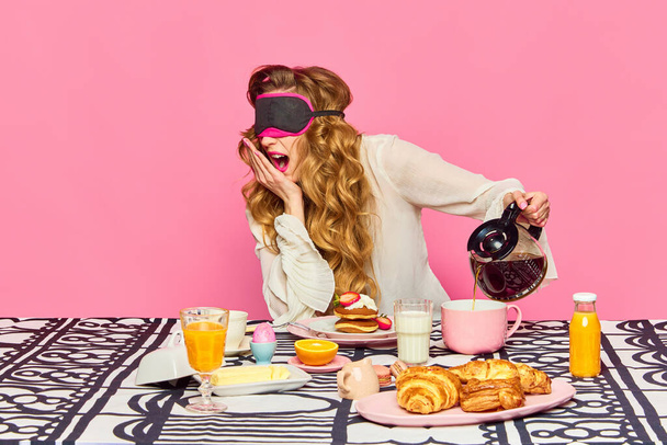 Fauler Morgen, französische Croissants, Kochen. Schläfriges schönes Mädchen mit lockigem Haar, das eine Schlafbrille trägt und Tee, Frühstück und Gähnen vor rosa Hintergrund zubereitet. Konzept von Morgen, Essen, Werbung - Foto, Bild