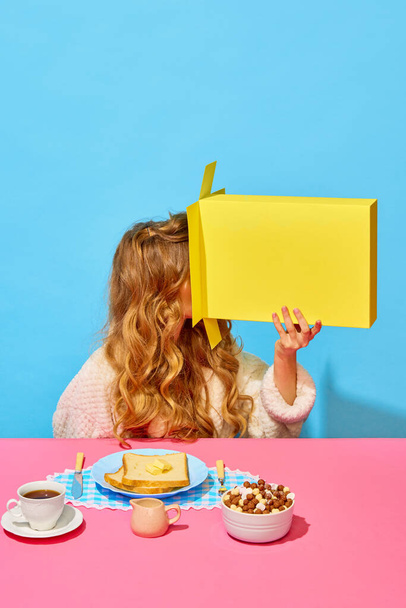 Yummy is over. Food pop art photography. Woman with long silk curly hair looking in empty cereal box over light blue pink background. Crazy morning, fun, joy and colorful minimalism concept - Foto, afbeelding