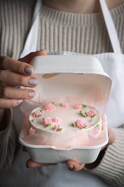 Mujer pastelera con delantal blanco y sosteniendo un pequeño pastel de bento decorado con flores de color rosa queso crema. Las manos de mujer sostienen el pastel de cumpleaños en una caja de regalo blanca. Pastel estilo coreano de moda - Foto, imagen