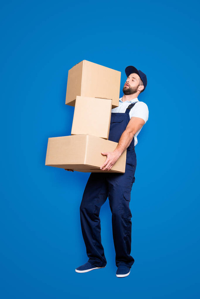 Full size body portrait of busy attractive deliver in blue uniform with stubble, having three heavy big boxes in arms, isolated on grey background, trying to withhold. - Valokuva, kuva