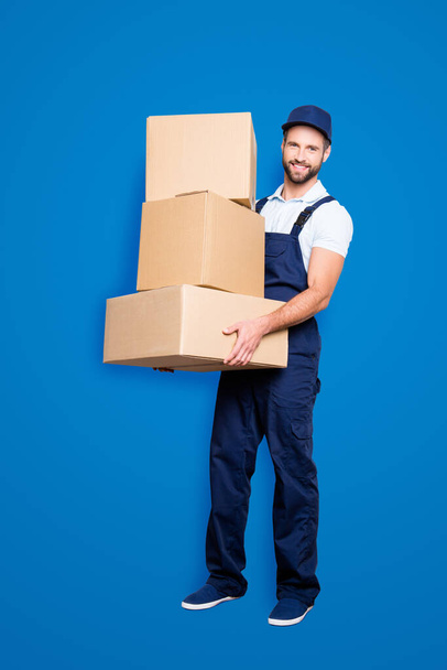 Full size fullbody portrait of attractive cheerful deliver in blue uniform with stubble, looking at camera having three big boxes in arms, isolated on grey background. - 写真・画像