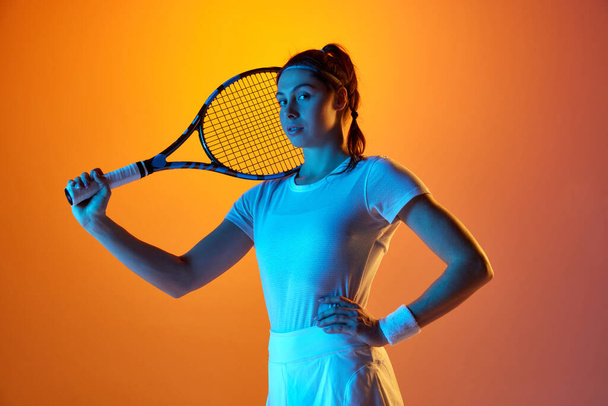 Young motivated woman, professional female tennis player in uniform posing with tennis racket against orange studio background in neon light. Concept of professional sport, movement, health, action - Фото, изображение