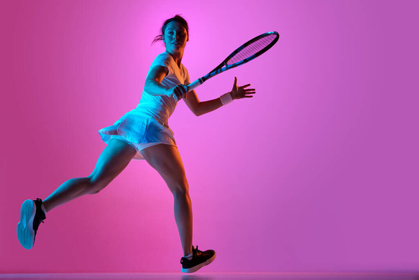 Full-length side view image of female tennis player in motion, wearing uniform, training against pink studio background in neon light. Concept of professional sport, movement, health, action. Ad - Foto, Imagen