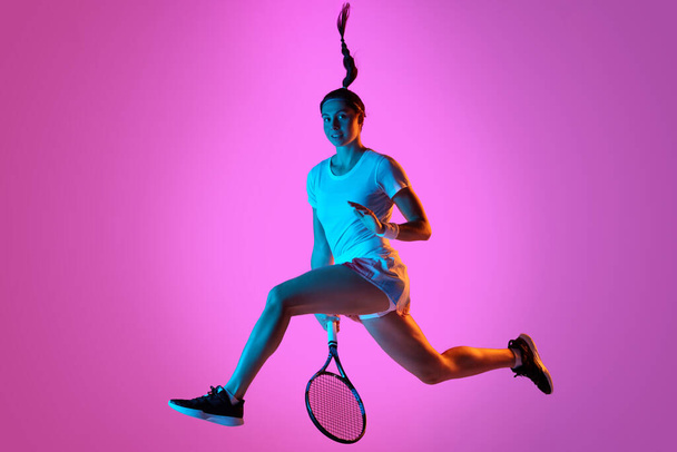 In a jump. Dynamic portrait of young athletic woman, tennis player in motion against pink studio background in neon light. Concept of professional sport, movement, health, action. Ad - Fotoğraf, Görsel