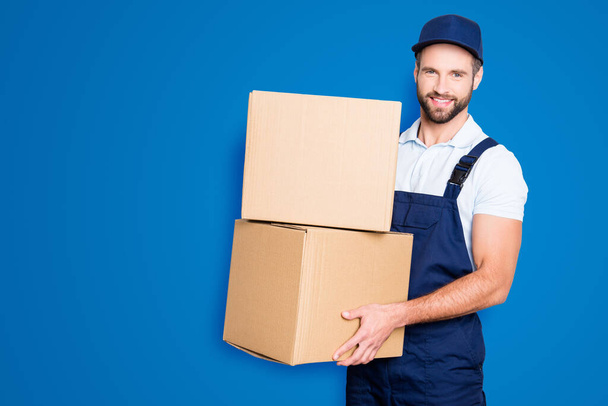 Portrait of attractive cheerful deliver in blue uniform with stubble looking at camera having two big boxes in hands, isolated on grey background. - Foto, Imagen