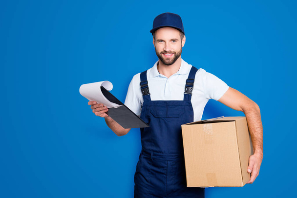 Portrait of cheerful positive deliver in blue uniform carrying having box and clipboard in hands, looking at camera, isolated on grey background. - Photo, Image