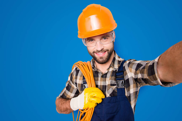 Self portrait of attractive joyful man in hardhat, overall, shirt, holding rolled cable on shoulder, blogger shooting selfie on front camera, isolated on grey background, having leisure. - Foto, afbeelding