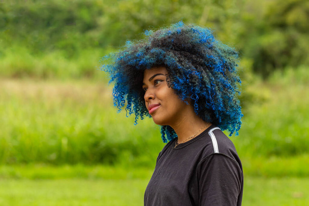 Foto van een jonge zwarte vrouw met afro haar, blauw geverfd, met een lichte glimlach op haar gezicht, in profiel met wazig park vegetatie op de achtergrond. - Foto, afbeelding