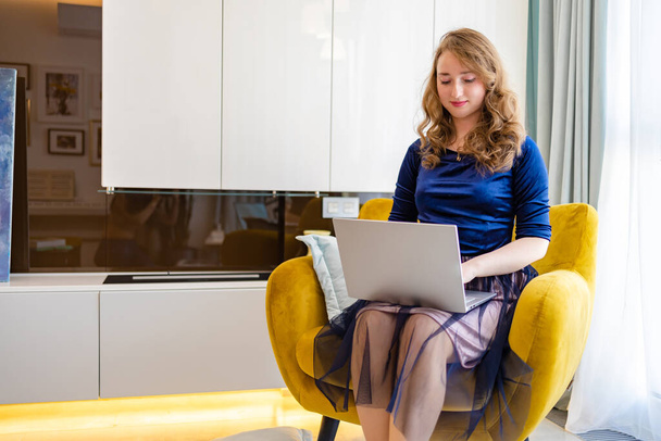 Bizneswoman w niebieskiej sukience pracuje z laptopem siedząc na kanapie w domu - Zdjęcie, obraz
