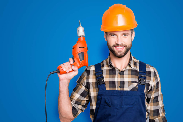 Portrait of handsome attractive repairer in protective hard hat, overalls, shirt with stubble showing having equipment in arm isolated on grey background. - Фото, зображення