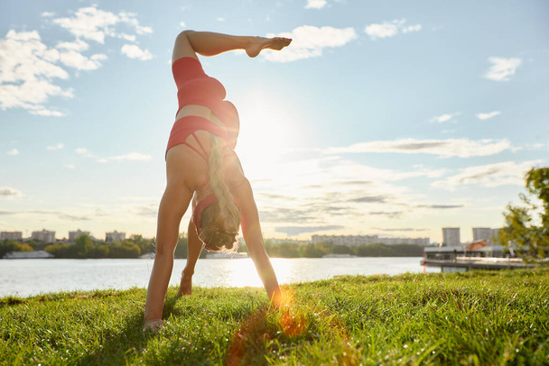 Mujer haciendo ejercicios de flujo de animales al aire libre al amanecer, en rayos de sol sobre hierba verde junto al río, postura escorpión. - Foto, imagen