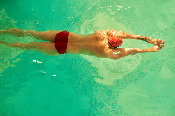 Un jeune homme nage rampant dans la piscine avec un bonnet rouge et des lunettes de natation, s'entraînant dans la piscine, se préparant pour la compétition, nageant, rampant devant, brasse, papillon. - Photo, image