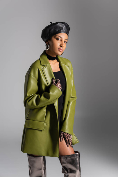 Fashionable african american model in leather beret and coat posing isolated on grey  - Foto, Imagen