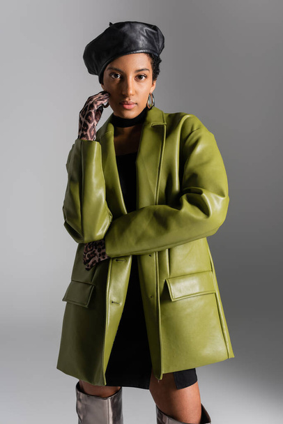 Модная африканская американка в перчатках с животной печатью и кожаным пальто, изолированным на сером  - Фото, изображение