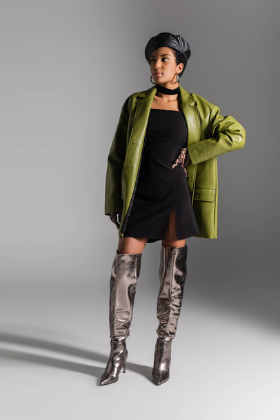 Mujer afroamericana de moda en botas brillantes y abrigo de cuero posando sobre fondo gris - Foto, Imagen