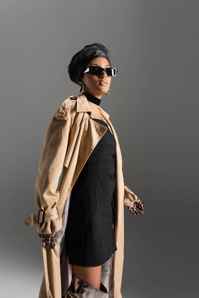 Elegante mujer afroamericana en gafas de sol y gabardina aislada en gris  - Foto, Imagen