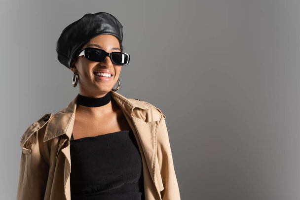 Smiling african american woman in beret and trench coat standing isolated on grey  - Foto, afbeelding