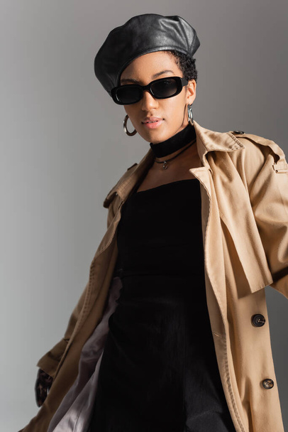 Trendy african american model in sunglasses and trench coat standing isolated on grey  - Фото, зображення