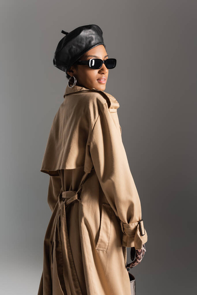 модна афро-американська модель в береті і траншеї пальто стоїть ізольовано на сірому
  - Фото, зображення