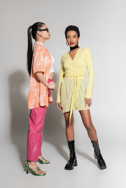 Full length of stylish interracial models in bright spring outfit posing on grey background  - Φωτογραφία, εικόνα