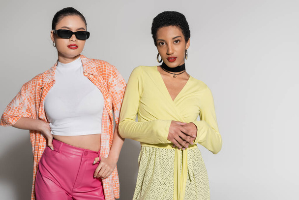 Модные азиатские и африканские американские модели в весеннем наряде, стоящие на сером фоне  - Фото, изображение