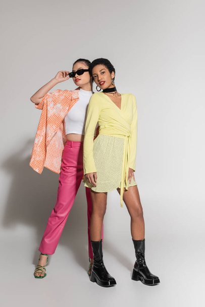 Full length of fashionable interracial models in colorful clothes standing on grey background  - Foto, Imagem