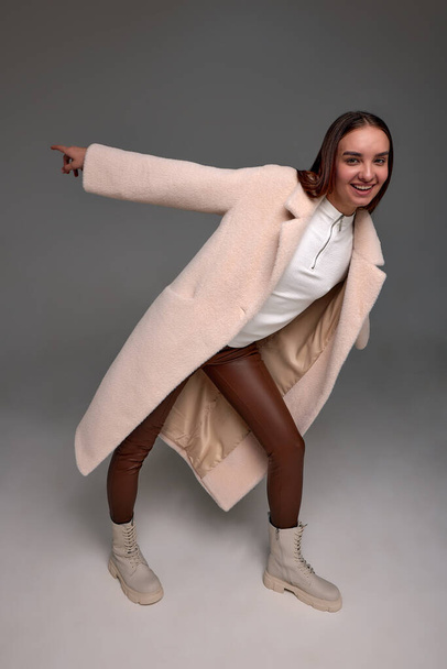 Retrato de una hermosa niña en abrigo de piel de oveja de moda y sombrero de punto de pie sobre el fondo de los abetos de invierno. Moda de invierno. Copiar espacio - Foto, imagen