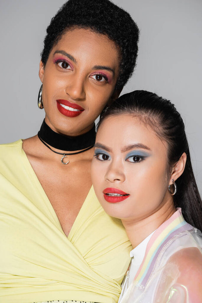 Positive multiethnic girlfriends with bright makeup looking at camera isolated on grey   - Φωτογραφία, εικόνα