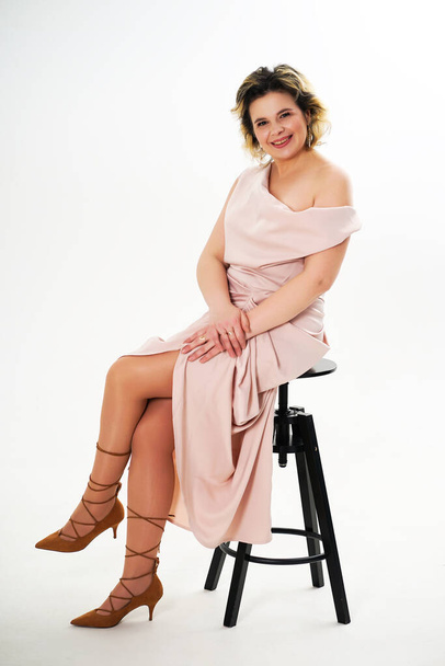 A woman in a beige dress sits on a black chair with her hands folded in front of her on a white background. - Foto, Bild