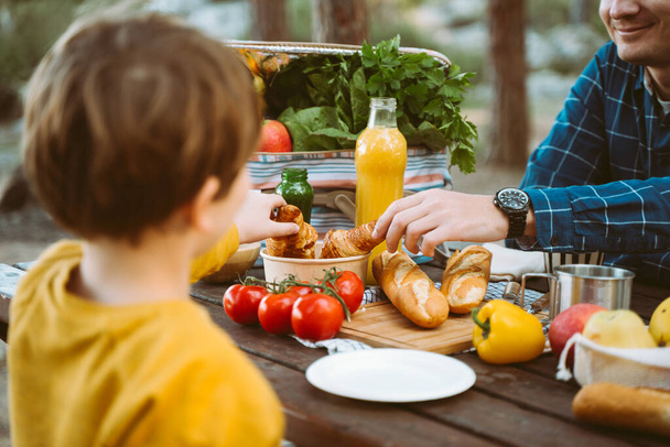 Isä isä koulupoika poika poika ottaa piknik metsässä leirintäalueella vihanneksia, mehua, kahvia, ja croissantit. Puinen laatikko tuoreita orgaanisia vihanneksia ympäröi leipää patonkeja - Valokuva, kuva