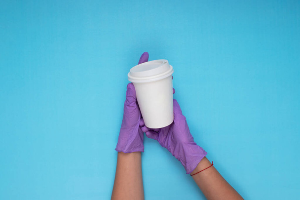 doctor in lilac colored gloves holding white cardboard coffee mug on light blue background - Photo, Image