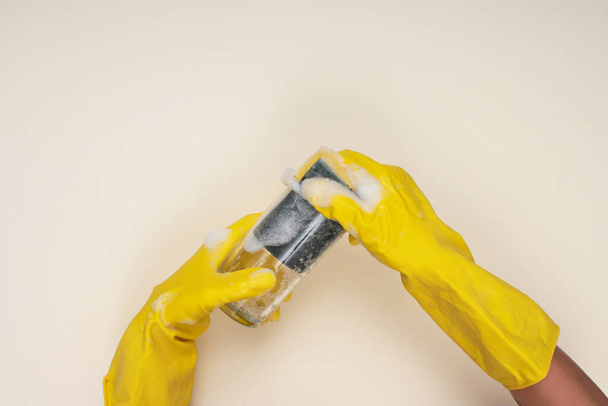 γυναίκα φορώντας κίτρινα γάντια καθαρισμού σε ανοιχτό χρώμα φόντο καθαρισμού γυάλινο κύπελλο με σφουγγάρι καθαρισμού - Φωτογραφία, εικόνα
