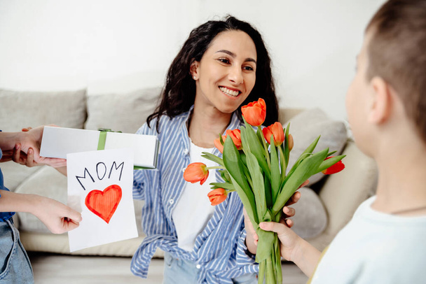 Arab Mom receives gifts from her children. The son holds out a bouquet of tulips and the daughter a handmade card and a gift. Everyone is in a bright room in a cozy homely atmosphere. - Фото, зображення