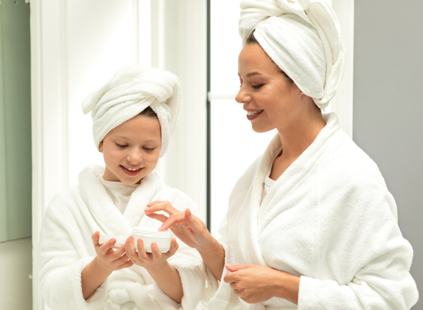 Smiling caucasian small daughter and little young mom in bathrobe and towel, have fun, apply cream, enjoy beauty care in bathroom. Relationship at home, skin hydration, ani aging procedures - Foto, Imagem